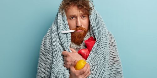 Redhead young man with bristle fights against flu and fever, holds lemon to enrich vitamins, has thermometer in mouth, trembles under blanket, isolated over blue background. Health problems.